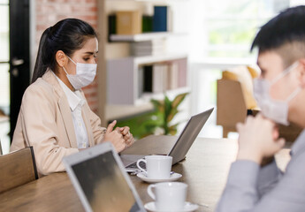 young Asian businesswoman wearing smart casual outfit sitting in the company office wearing face mask looking on smartphone and laptop while working. Selective focus on female worker