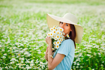 A young brunette with a hat on her head in a blue dress holds a bouquet of flowers in her hands and sits on a chamomile field, enjoying the aroma of flowers.