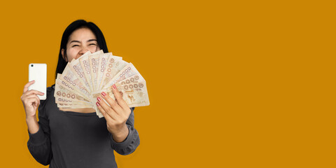 happy Asian woman showing Thai baht money another hand holding smart phone over orange banner background - obrazy, fototapety, plakaty