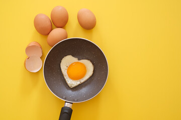 Heart shape fried egg in the pan with egg shell and chicken eggs on yellow background. top view. healthy food  concept. Copy space for text 