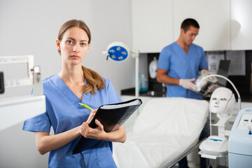Obraz premium Young polite female cosmetologist meeting patient in office of aesthetic medicine, filling out medical form .