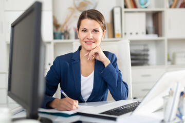 Portrait of successful busy female entrepreneur sitting at office desk with papers and laptop - obrazy, fototapety, plakaty