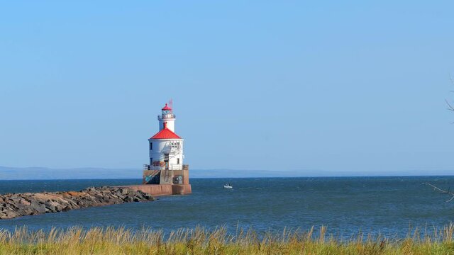 Lighthouse with red roof at end of pier and fishing boat on the blue water, on a sunny day, as the nearby grass sways gently in the breeze. Zoom in.