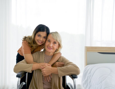 Happy mix skin family living together, adopted asian daughter cuddle caucasian mother sitting on a wheelchair with smiles. Concept of love and sharing time of family, mother or daughter in law.