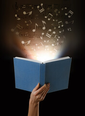 Female hand holding open book with flying music notes on dark background