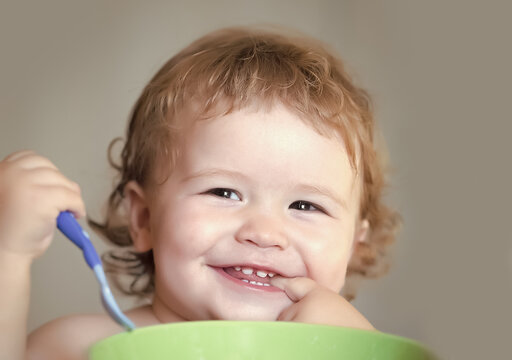 Baby eating. Portrait of funny little smiling boy with blonde curly hair and round cheecks eating from green plate holding spoon closeup.