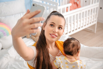 Young mother with little baby in sling taking selfie at home