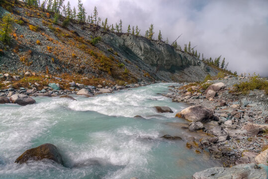 The mountain river flows along the steep beren. Low clouds float above the ground.