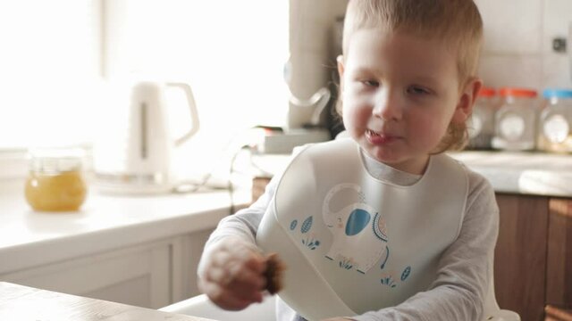 A boy in a bib and a highchair sits at the kitchen table. Eats a healthy coffee and enjoys it.