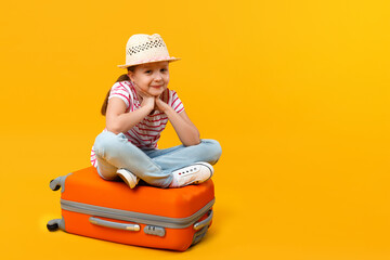 The child sits on a suitcase and holds his cheeks with his palms on a yellow background. Little...