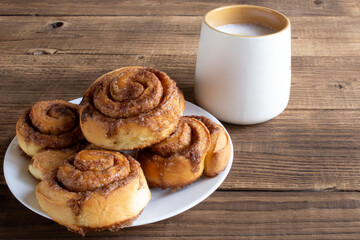 Freshly baked cinnamon roll with spices and cocoa filling and coffee or cappuccino on white serving plate on wooden background. Cinnabon buns. Swedish breakfast.