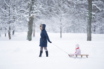 Happy young adult mother pulling wooden sledge with baby girl on snowy park road in deep fresh snow. Enjoying white winter day. Spending time together in weekend. Side view.