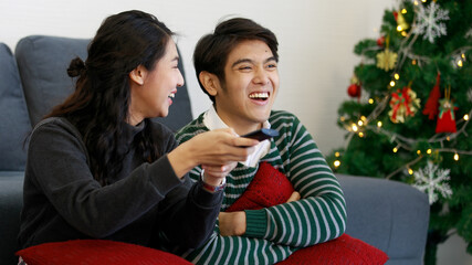 Portrait of young adult Asian lover couple in a sweatshirt resting on the sofa in the living room at home, arguing and fighting to get television remote control to select the movie to watch together