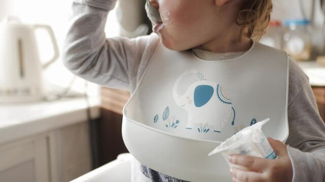 The child eats cottage cheese with appetite, licking a spoon of yogurt from a jar, his face is dirty. A boy in a bib and a highchair sits at the kitchen table.