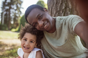 Smiling dark-skinned man making selfie with his little daughter in the park