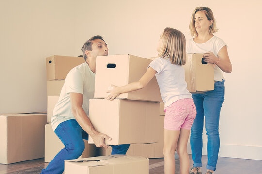 Young family carrying boxes into new home or apartment. Cute daughter helping father with belongings. Blonde mother in jeans standing and holding box. Mortgage, relocation and moving day concept