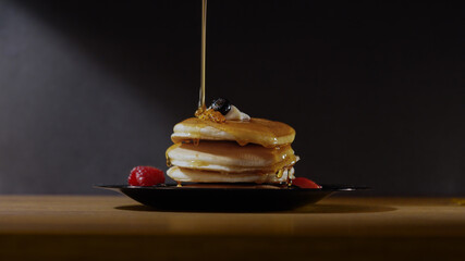 Golden Maple syrup poured onto a stack of delicious pancakes with fruit over a black background.