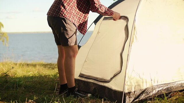 A tired tourist comes to his tent standing on the shore of the sea, river, lake. He enters the tent and closes the door. Tourist weekend on the shore of sea, lake, river.