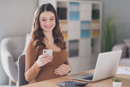 Photo of young pregnant manager sit table hold phone look screen wear brown shirt workstation office indoors