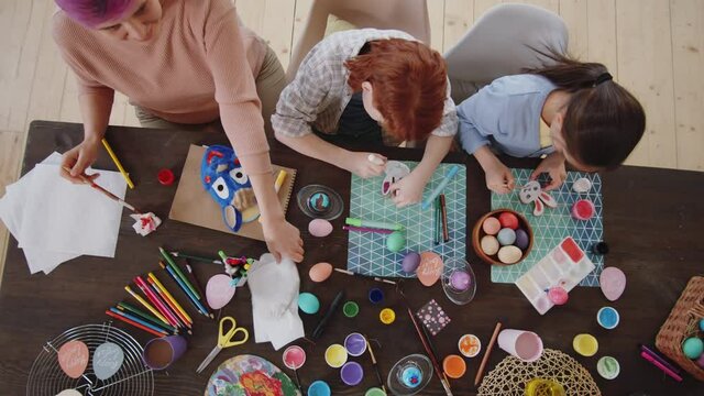 Top down shot of little daughter, son and mother sitting at table with paints and craft supplies and making decorative Easter toys