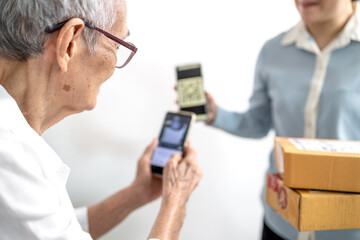 Asian senior woman receiving parcel post box from delivery service,paying deliver with smartphone to scan QR code payment purchase,pay without money,contactless cashless society,old elderly technology