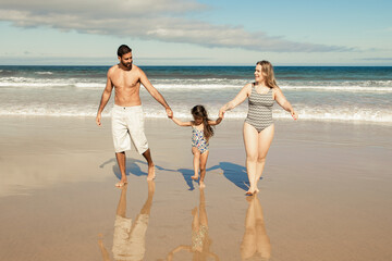 Parents couple and little daughter wearing swimsuits, walking on golden sand from sea. Girl holding parents hands. Front view. Family outdoor activities concept