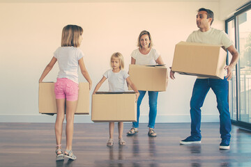 Fototapeta na wymiar Caucasian parents and two girls holding carton boxes and standing in empty living room. Lovely daughters helping during relocation in new apartment. Mortgage, property and moving day concept