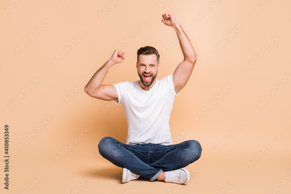 Sticker Portrait of nice cheery guy sitting on floor rejoicing victory triumph isolated over beige color background