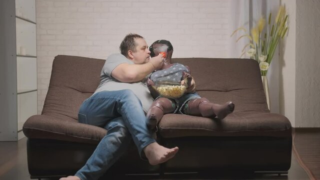 adult man watches TV and eats popcorn in company of doll while sitting on couch in room.