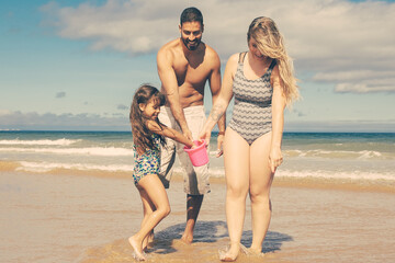 Happy family with little girl walking ankle deep in sea water and wet sand, picking shells into bucket. Bright blue sea with waves and white foam in background. Family summer holidays concept
