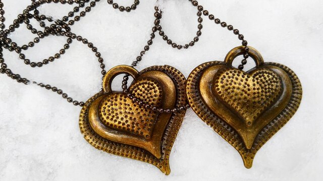 Two gold hearts on white snow connected by metal chain. Love friendship romantic couple concept with gold heart top view. Gold vintage trinket or pendant in heart shape lovers background or card image