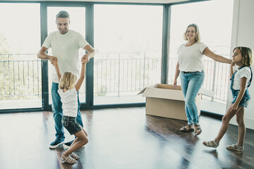 Happy mom, dad and kids having fun during removal. Cheerful father playing with cute girl in empty room and smiling. Mother and sister looking at them. Mortgage, relocation and moving day concept