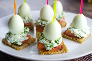 canapes with salmon and quail eggs on skewers, delicious small sandwiches, bread with cream cheese and fish on a white plate