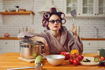 Funny angry young housewife in bad mood cooking lunch and waiting for husband at home. Woman in bathrobe, curlers, thug life 8 bit pixel sunglasses holding spatula sitting at kitchen table like a boss