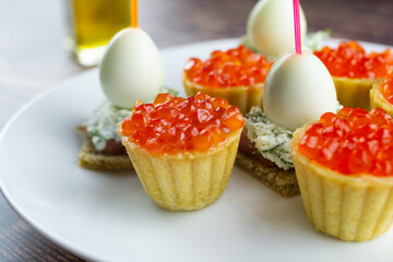 canapes with red caviar, salmon and quail eggs on a white plate, tartlets, small appetizing sandwiches on skewers, bread with cream cheese on a wooden background