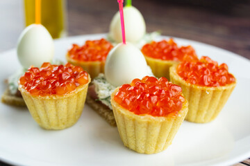 canapes with red caviar, salmon and quail eggs on a white plate, tartlets, small appetizing sandwiches on skewers, bread with cream cheese on a wooden background
