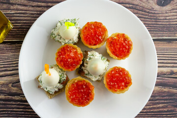 canapes with red caviar, salmon and quail eggs on a white plate, tartlets, small appetizing sandwiches on skewers, bread with cream cheese on a wooden background