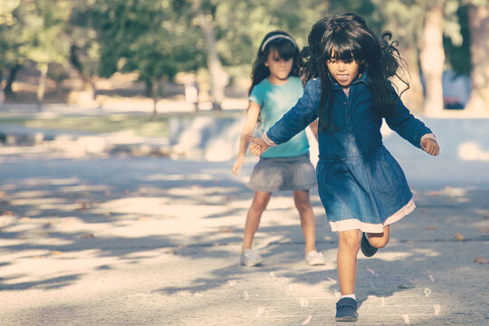 Two excited black haired little girls playing hopscotch in city park. Full length, copy space. Childhood concept