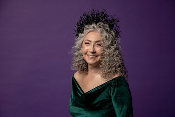 Studio portrait of an elderly woman 60-65 years old in a beautiful long green dress with a crown on her head on a colored background. Concept: stylish pensioners of model appearance, active life.