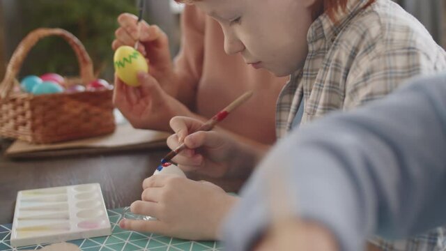 Close up view of little boy drawing on egg with paintbrush while making Easter decorations with family at home