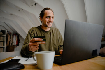Caucasian male student smiling while sitting at desk successfully affording monthly rent enjoying hot coffee