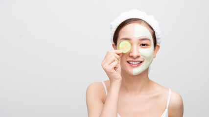 Beautiful young Asian woman with clay mask face holding cucumber piece over white background.