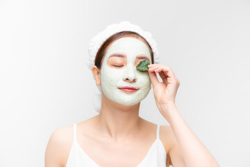 smiling girl with a white towel on her head applied a clay moisturizing mask while holding a leaf over white background.