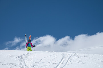 A young woman in ski equipment stands in the middle of a snowy field in the mountains against a background of blue sky and cloudy mountains. Freeride backcountry