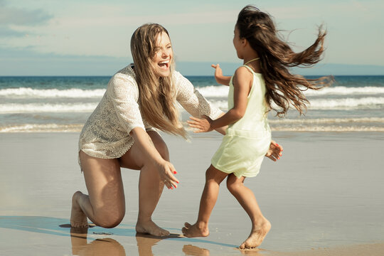 Joyful mom opening arms for catching and hugging little kid. Girl running to mother on beach at sea. Parenting and outdoor activities concept