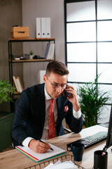 Young businessman using computer in his office. Handsome man talking to the phone at his workplace.