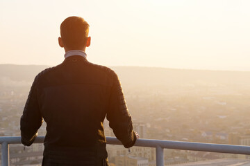 Muscular man in black jacket holds hands on grey metal handrails and admires sunrise over cityscape and hills silhouettes backside view