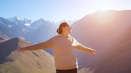 Happy woman wearing sunglasses and pink hoodie enjoys bright sunshine rays standing against amazing mountain range peaks