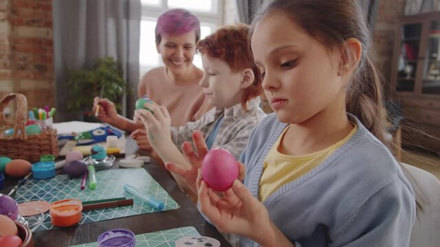 Beautiful little girl applying paint to Easter egg with finger while doing crafts with mother and brother at home
