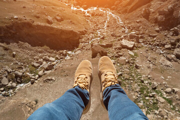 Man's legs over a cliff and a mountain river, sunlight.